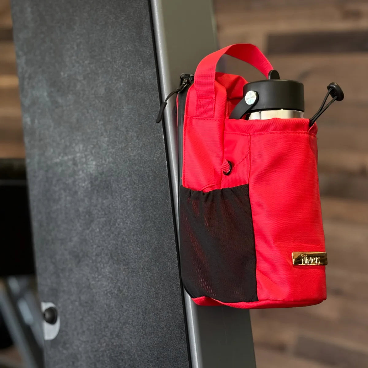 Red Nimbis magnetic gym bag attached to a metal gym machine, holding a bottle with a blurred background.